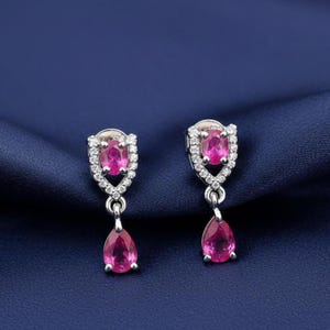 May include: A pair of silver-toned drop earrings featuring a central oval-cut pink gemstone surrounded by small clear stones, with a dangling pear-shaped pink gemstone suspended from the base of each earring setting.