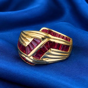 Puede incluir: Un anillo ancho de oro con múltiples rubíes rojos de talla baguette dispuestos en un patrón geométrico. La banda de oro amarillo pulido tiene secciones curvas y lisas que contrastan con la disposición lineal de las piedras preciosas rojas.