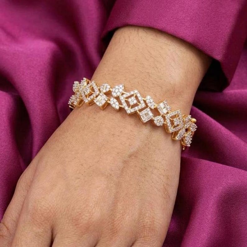 May include: A gold bracelet with a geometric pattern, incorporating square and rectangular elements, and set with small, sparkling stones. The bracelet is displayed on a wrist against a deep purple fabric.