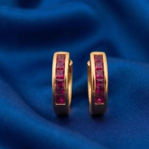May include: A pair of gold hoop earrings featuring a channel setting with five square-cut red rubies embedded in the front of each hoop. The earrings have a polished gold finish and a classic circular design, showcasing vibrant red gemstones.