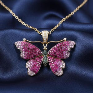 May include: A gold-toned butterfly pendant necklace with a chain. The butterfly wings are encrusted with pink and white gemstones, with a green gemstone body. The necklace is displayed on a dark blue satin fabric.