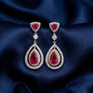 May include: A pair of elegant drop earrings with a pear-shaped design. Each earring features a vibrant red gemstone at the center, surrounded by a halo of sparkling white stones. The earrings have a triangular top and are set against a dark blue background.