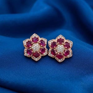 May include: A pair of flower-shaped stud earrings crafted from rose-toned metal. Each earring features six oval-cut deep pink rubies arranged as petals surrounding a central cluster of small, clear, round-cut white stones set in a circular pattern.