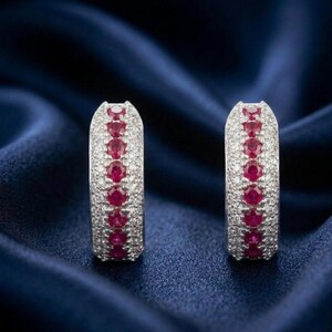 May include: A pair of silver hoop earrings. Each earring is encrusted with small, clear stones and a row of vibrant red gemstones. The earrings are set against a dark blue, silky background.