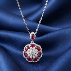 Puede incluir: Un collar de tono plateado con un colgante en forma de flor. El colgante presenta un grupo central de pequeñas piedras claras rodeado por seis piedras rojas de corte ovalado, con una piedra roja adicional de corte marquesa en la parte superior.