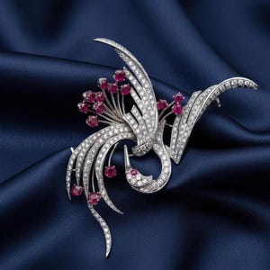 May include: A silver-coloured brooch depicting a bird with open wings, embellished with many small, sparkling clear stones and clusters of red gemstones. The brooch is set against a deep blue satin background.