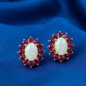 Könnte beinhalten: Ein Paar Ohrstecker aus Gold mit einem zentralen ovalen weißen Opal, der von einem Kranz aus kleinen, runden, tiefroten Rubinen umgeben ist. Die Steine sind blumenartig angeordnet und bilden einen lebendigen Kontrast zwischen dem schillernden Opal und den roten Edelsteinen.