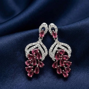 May include: A pair of silver-toned drop earrings with a leaf-like design. The earrings feature a cluster of teardrop-shaped, ruby-colored gemstones and are encrusted with clear, sparkling crystals. The earrings are displayed on a dark blue satin background.