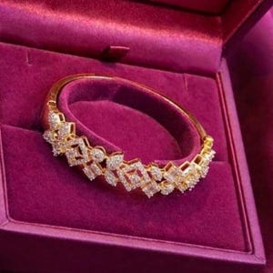 May include: A gold bracelet with a diamond-like pattern, displayed in a magenta velvet jewellery box. The bracelet has a series of geometric shapes set with clear, sparkling stones. The box is open, showcasing the bracelet's intricate design.