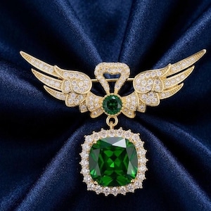 May include: A gold-toned brooch with a wing design, encrusted with small, clear crystals. The brooch features a large, square, emerald-green gemstone pendant, surrounded by smaller crystals. The wings are spread, and the brooch is set against a dark blue background.