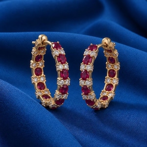 May include: A pair of gold hoop earrings featuring alternating round-cut red rubies and clear white diamonds set in a continuous circular pattern. The earrings have a hinged closure mechanism and a polished gold finish throughout the metal frame.