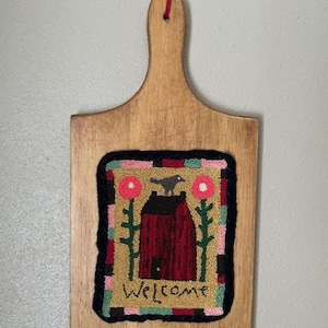 May include: A wooden cutting board with a hand-stitched welcome sign featuring a red house, two pink flowers, and a black bird. The sign is surrounded by a colorful border.