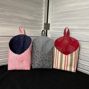 May include: Three fabric hanging storage pockets. The first is pink with white stripes and a navy blue polka dot interior. The second is gray with black polka dots. The third is red with a white and green striped pattern.