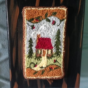 May include: A framed, rectangular, hand-stitched wall hanging featuring a rustic cabin scene. The cabin has a red roof, with green trees and a white background. The frame is dark brown with a wood grain appearance.