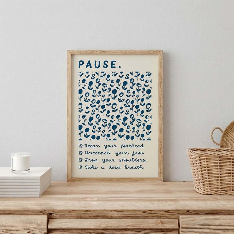 May include: A rectangular wall art print in a light wood frame featuring the word PAUSE in dark blue capital letters. Below, a pattern of dark blue flowers on a cream background, followed by the text: Relax your forehead, Unclench your jaw, Drop your shoulders, Take a deep breath.