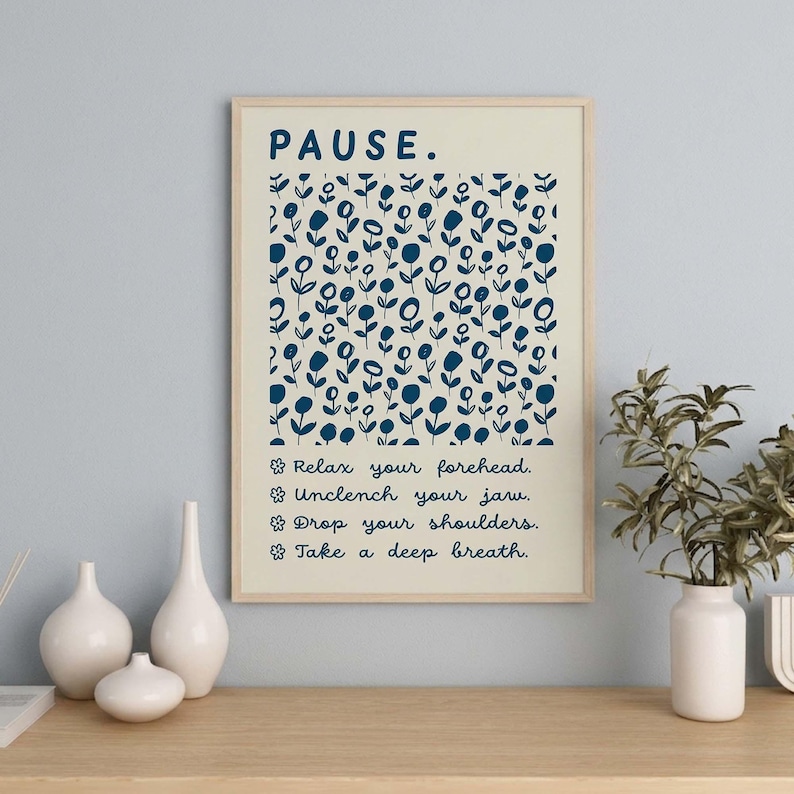 May include: A rectangular wall art print in a light wood frame featuring a cream background with a dark blue floral pattern. The text reads: PAUSE. Relax your forehead. Unclench your jaw. Drop your shoulders. Take a deep breath. Each line is preceded by a small flower icon.