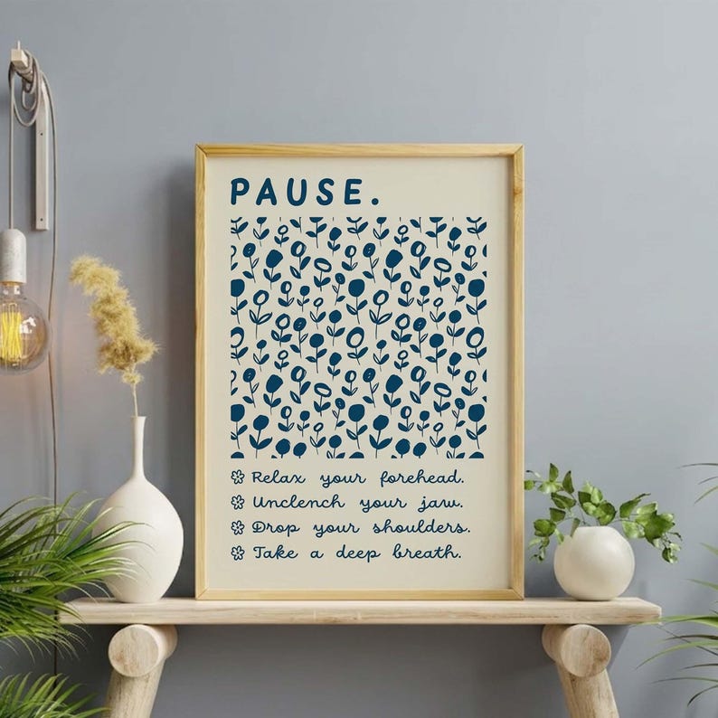 May include: A rectangular wall art print in a light wood frame featuring the word PAUSE in dark blue, a repeating pattern of dark blue line-drawn flowers, and four bulleted lines of text: Relax your forehead, Unclench your jaw, Drop your shoulders, Take a deep breath.