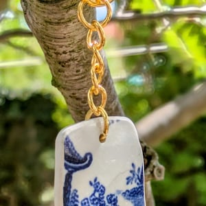 May include: A gold-coloured keyring with a rectangular, white ceramic pendant. The pendant features a blue and white landscape design. The keyring is hanging from a tree branch against a blurred green background.