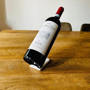 Könnte beinhalten: Eine Flasche Rotwein mit weißem Etikett, die auf einem weißen Weinhalter steht. Die Flasche ist angewinkelt, wodurch der Text und das Design des Etiketts sichtbar werden. Die Szene spielt auf einem Holztisch.