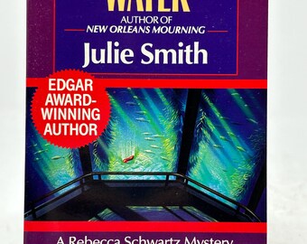 May include: A paperback book titled "Dead in the Water" by Julie Smith. The cover is predominantly purple with yellow and white text. The book features an image of an aquarium interior with fish. The cover also includes the text "Edgar Award-Winning Author."