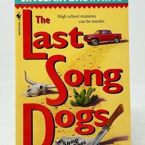 May include: A paperback book titled "The Last Song Dogs" by Sinclair Browning. The cover features a red pickup truck, a steer skull, a cactus, and a knife. The background is yellow with red text.