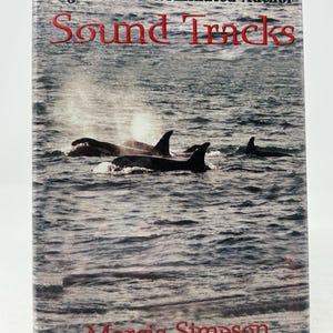 Könnte beinhalten: Buchcover von "Sound Tracks" von Marcia Simpson, mit Orcas im Meer. Titel und Autorenname sind rot. Der Text "Edgar Award Nominated Author" befindet sich oben auf dem Cover.