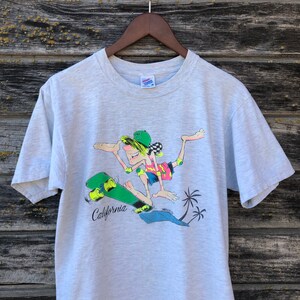 May include: A light grey t-shirt with a cartoon skater. The skater is wearing a green hat, pink and yellow clothing, and knee pads. The skateboard is green with yellow wheels, and the word "California" is printed on the board.