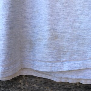 May include: Close-up of a light grey t-shirt. The fabric has a textured appearance with subtle colour variations. The bottom hem is visible, featuring a double-stitched seam in a lighter shade of grey. The shirt appears to be made of a soft, comfortable material.