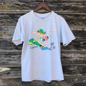 May include: Light grey t-shirt with a cartoon skater in a colourful outfit on a green skateboard. The word "California" is printed in black below the skateboard. Two palm trees are also in the design. The shirt is hanging on a wooden hanger.