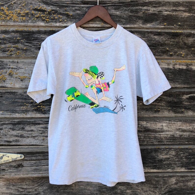 May include: Light grey t-shirt with a cartoon skater in a colourful outfit on a green skateboard. The word "California" is printed in black below the skateboard. Two palm trees are also in the design. The shirt is hanging on a wooden hanger.