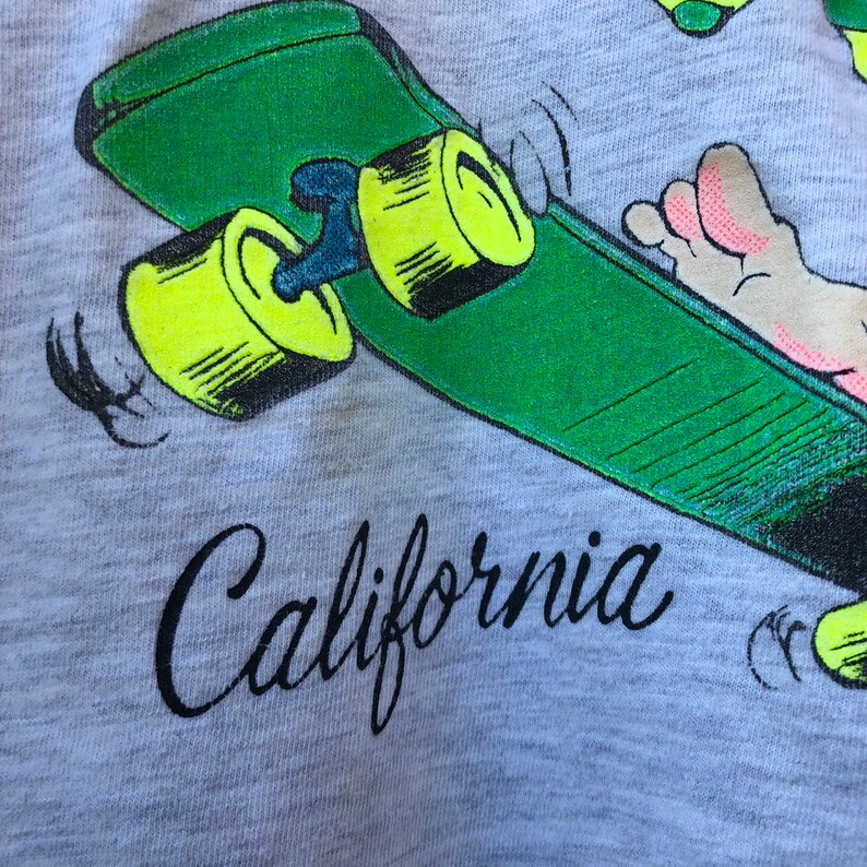 May include: A grey t-shirt with a green skateboard design and yellow wheels. The word "California" is written in black script. A cartoon foot is also visible on the skateboard.