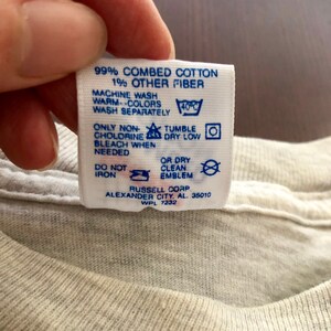 May include: A white clothing label with blue text and symbols, detailing washing instructions. It advises machine washing at 40&deg;C, tumble drying on low, and avoiding ironing. The label also features the brand name "RUSSELL CORP" and the location "ALEXANDER CITY, AL. 35010".