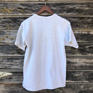 May include: A grey t-shirt hanging on a wooden hanger. The short-sleeved shirt is made of a soft material and has a crew neck. The background is a weathered wooden surface.