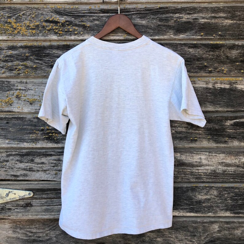 May include: A grey t-shirt hanging on a wooden hanger. The short-sleeved shirt is made of a soft material and has a crew neck. The background is a weathered wooden surface.