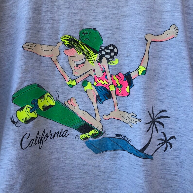 May include: A light grey t-shirt with a cartoon skater in action. The skater is wearing a green cap with "WAY COOL" text, a pink and yellow top, and blue shorts. The skateboard is green with yellow wheels, and the word "California" is printed below.