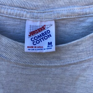 May include: Close-up of a light grey t-shirt with a label. The label reads "JERZEES COMBED COTTON" in blue and white, with a red logo. It also states "MADE IN U.S.A." and the size "M 38-40".