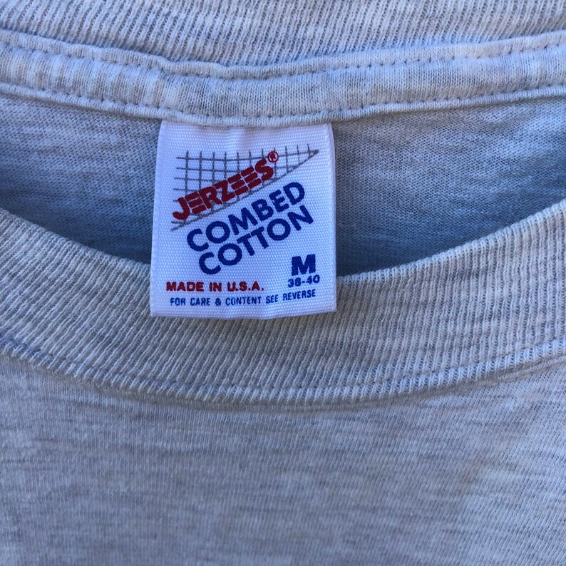 May include: Close-up of a light grey t-shirt with a label. The label reads "JERZEES COMBED COTTON" in blue and white, with a red logo. It also states "MADE IN U.S.A." and the size "M 38-40".