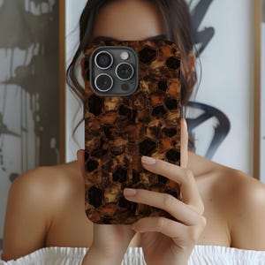 May include: A brown and gold phone case with a geometric pattern. The case has a dark camera module with three lenses. The case is held in front of a person.