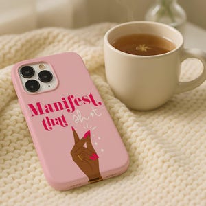 以下が含まれることがあります： ピンク色の携帯電話ケース。太字のピンク色のフォントで「Manifest that sh*t」と書かれています。ケースには、金の指輪とピンクの爪を持つ手のグラフィックが描かれています。携帯電話ケースは、クリーム色のニットブランケットの上に置かれています。