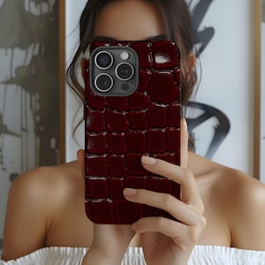 Può includere: Una custodia per telefono bordeaux con un motivo strutturato a pelle di coccodrillo. La custodia ha un modulo fotocamera con tre obiettivi e un flash. La custodia del telefono è tenuta davanti al viso di una persona.