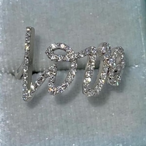 May include: A silver ring spelling "love" in a cursive font, studded with small, sparkling gemstones. The ring is displayed in a light blue jewellery box.