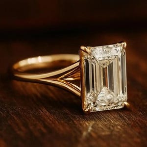 Puede incluir: Un anillo de compromiso de oro con un gran diamante rectangular con talla esmeralda. La banda del anillo tiene un diseño de vástago dividido. El diamante es claro y refleja la luz, sobre un fondo de madera oscura.