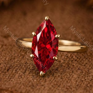May include: A gold ring featuring a large, marquise-cut red gemstone held by six prongs. The text MysticLabGrownJewlry is visible across the image. The band is a smooth, polished gold metal, and the stone displays a deep, vibrant red color.