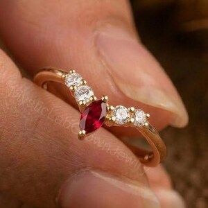 Pode incluir: Um anel dourado com um design em forma de V. O anel apresenta uma pedra preciosa vermelha escura com corte marquesa no centro, ladeada por pequenas pedras preciosas redondas e claras. O anel é segurado por uma mão.