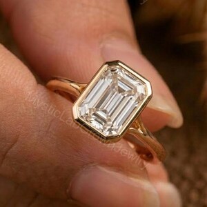 May include: A gold ring with a large, rectangular, emerald-cut diamond. The diamond is set in a bezel setting, and the ring has a slender band. The ring is held by a person.
