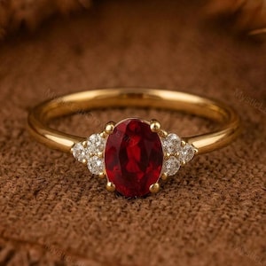 May include: A gold ring with a central oval-cut, deep red gemstone. Either side of the main stone are clusters of smaller, clear gemstones. The ring has a simple, polished gold band. The ring is displayed on a textured brown surface.