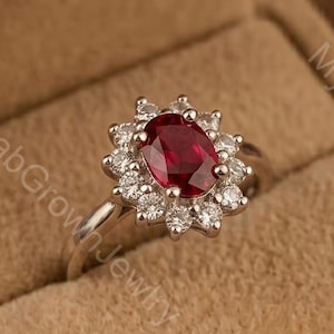 May include: A silver-toned ring featuring a central oval-cut deep red gemstone surrounded by a halo of small, round, clear stones. The ring is presented inside a tan, velvet-lined jewellery box with the text MysticLabGrownJewlry visible.