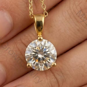 May include: A gold pendant necklace featuring a large, round, clear gemstone. The gemstone is set in a gold bezel and hangs from a delicate gold chain. The necklace is displayed against a neutral background.
