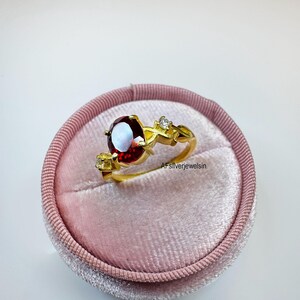 Può includere: Un anello color oro con una grande pietra preziosa rosso scuro dal taglio ovale come elemento centrale, affiancata da due pietre d'accento più piccole, chiare e dal taglio rotondo su una fascia delicata e testurizzata. Il testo AFsilverjewelsin è visibile.