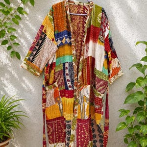 May include: A long-sleeved robe featuring a patchwork design made of various fabric scraps in shades of red, yellow, orange, green, and blue. The garment has an open front, a tie belt, and visible white stitching throughout the quilted material.